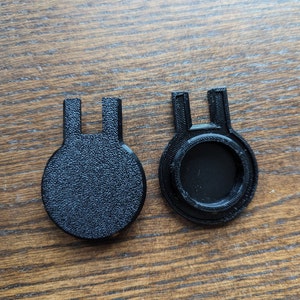 Op de afbeelding: Twee zwarte plastic afdekkingen met een U-vormige opening. Een afdekking heeft een rond gat in het midden.