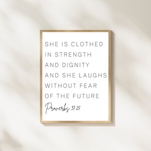 May include: A framed print with a white background and black text that reads "She is clothed in strength and dignity and she laughs without fear of the future. Proverbs 31:25".