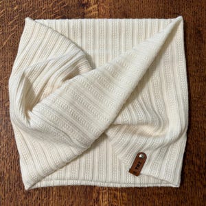 May include: Cream-colored ribbed knit headband with a twisted design. The headband features a small, brown leather tag with embossed text. The fabric has a textured, vertical stripe pattern.