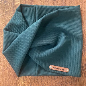 May include: A dark teal-colored headband with a textured, quilted pattern. The headband is twisted at the front and features a small, rectangular leather tag with the text "MERLE & TRI" attached with two silver rivets.