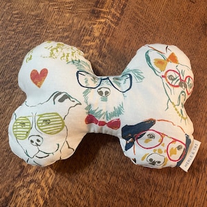 May include: A dog bone-shaped dog toy with a white background and colorful illustrations of dogs wearing glasses and bow ties. The toy has a red heart and the text "MERLE & TRI" on a small tag.