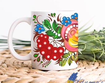 Op de afbeelding: Witte keramische mok met een levendig bloemmotief in rood, roze, blauw en geel. De mok heeft een gebogen handvat en is versierd in een traditionele volkskunststijl. De mok staat op een geweven oppervlak.