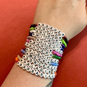 Puede incluir: Una pila de pulseras de cuentas de colores con cuentas blancas que deletrean diferentes palabras, incluyendo "Rogue", "Druid", "Wizard", "Bard", "Ranger", "Fighter", "Monk", "Sorcerer", "Paladin", y "Barbarian".