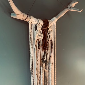 May include: A white and brown macrame wall hanging with wooden branches, white beads, and brown leather cords.
