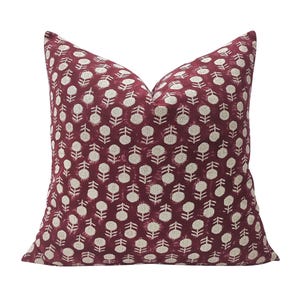 May include: A square throw cushion featuring a deep burgundy red background with a repeating pattern of small, off-white stylised floral motifs. The design consists of circular flower heads on short stems arranged in a consistent grid-like layout.