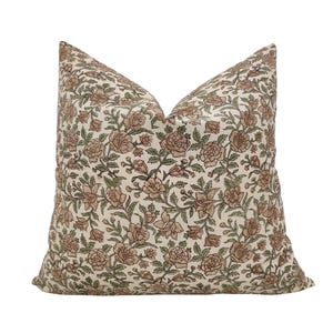 Puede incluir: Un cojín decorativo cuadrado con un estampado floral. La tela crema está cubierta con un patrón repetido de flores y hojas marrones y verdes. El cojín está diseñado para uso decorativo y es adecuado para una sala de estar o un dormitorio.