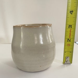 May include: A small, white ceramic vase with a rounded shape and a slightly flared rim. The vase has a subtle, off-white glaze and a smooth, matte finish.