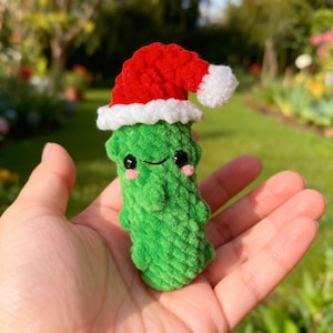 May include: A handmade green crochet pickle wearing a red and white Santa hat. The pickle has black eyes and pink cheeks, giving it a cheerful appearance. The plush toy is held in a hand, with a blurred green background.