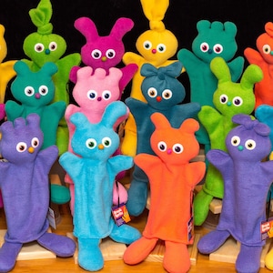 May include: A collection of colorful hand puppets, including a yellow, green, pink, blue, purple, orange, and teal puppet. The puppets have large eyes and are made of soft fabric.