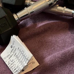 Könnte beinhalten: Ein kleines, gefaltetes Notizbuch mit handgeschriebenen Trainingsnotizen. Das Notizbuch liegt auf einem lila Handtuch mit zwei schwarzen und silbernen Hanteln darauf. Das Notizbuch listet Übungen und Sätze mit Gewichten und Wiederholungen auf. Der Text im Notizbuch lautet: "easy I miles", "Pilates 45min", "weight", "reps", "Rows-Bat", "70", "4", "Lat Rul domes", "100", "4", "Cable Fig", "10", "3", "Bar Curl", "70", "4", "Prewadex Corl", "25", "4", "Baby wil", "25", "4", "Calf Ra 100", "5".
