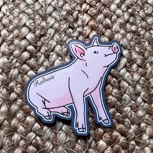 May include: A pink pig sticker with the word "Radiant" written on it. The pig is sitting on a woven straw background.