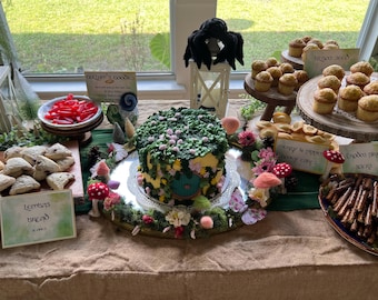 May include: A whimsical dessert table featuring a fairy-themed cake with green frosting and floral accents. Other treats include cupcakes, scones, and red candies, all arranged with decorative mushrooms and signage.