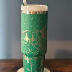 May include: A green stainless steel tumbler with a silver lid and a straw. The tumbler has a design of mountains, trees, and bears etched into the side.