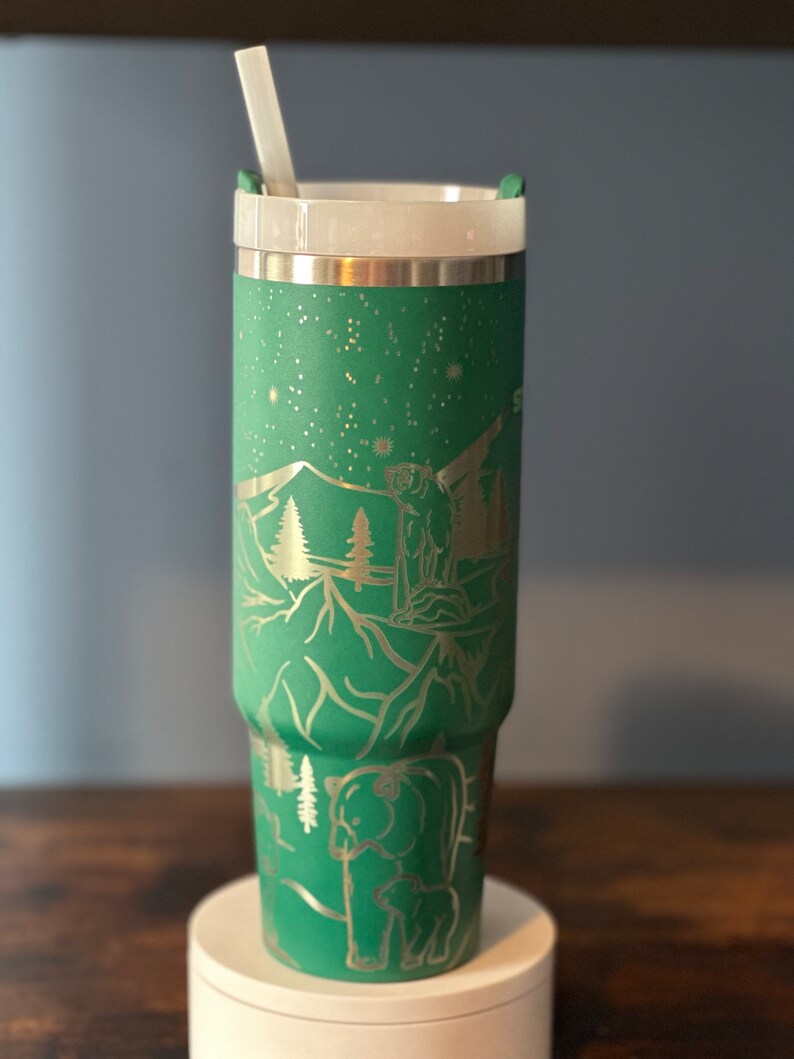 May include: A green stainless steel tumbler with a silver lid and a straw. The tumbler has a design of mountains, trees, and bears etched into the side.