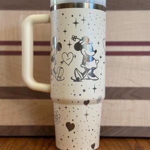 May include: A white stainless steel travel mug with a black handle. The mug has a silver design of Mickey Mouse and Minnie Mouse with hearts and stars.