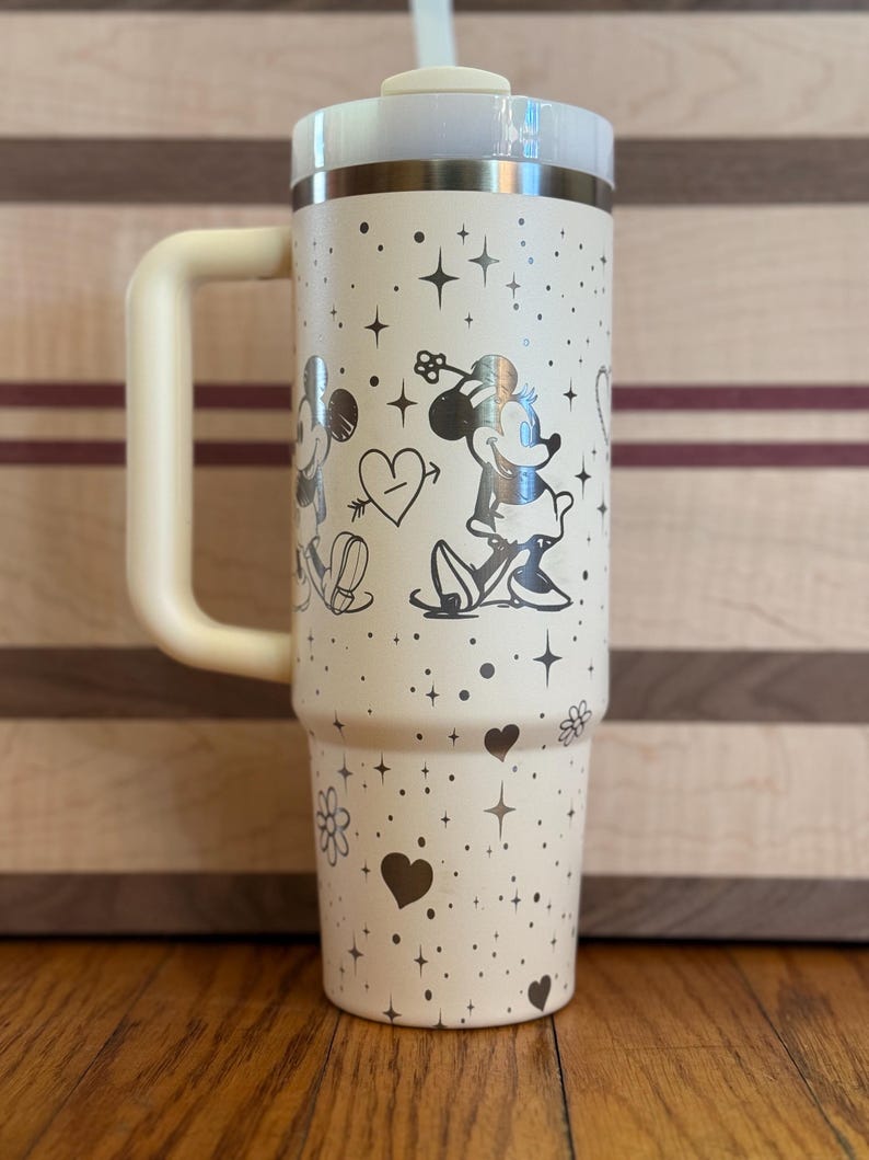 May include: A white stainless steel travel mug with a black handle. The mug has a silver design of Mickey Mouse and Minnie Mouse with hearts and stars.