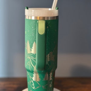 May include: A green stainless steel tumbler with a white lid and a clear straw. The tumbler has a handle and a design of trees and mountains on it.