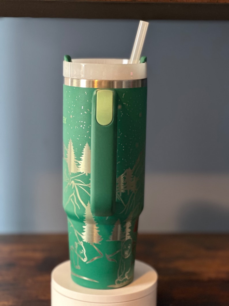 May include: A green stainless steel tumbler with a white lid and a clear straw. The tumbler has a handle and a design of trees and mountains on it.