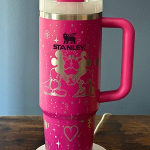 May include: Pink stainless steel travel mug with a black and white Mickey Mouse and Minnie Mouse design. The mug has a black Stanley logo and a pink handle.