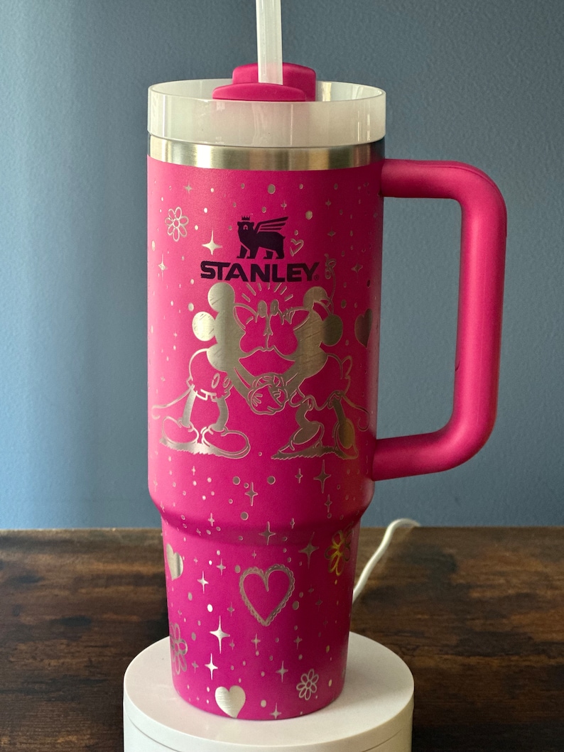 May include: Pink stainless steel travel mug with a black and white Mickey Mouse and Minnie Mouse design. The mug has a black Stanley logo and a pink handle.