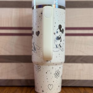 May include: White stainless steel tumbler with a black and white Mickey Mouse and Minnie Mouse design. The tumbler has a black lid and a black handle.