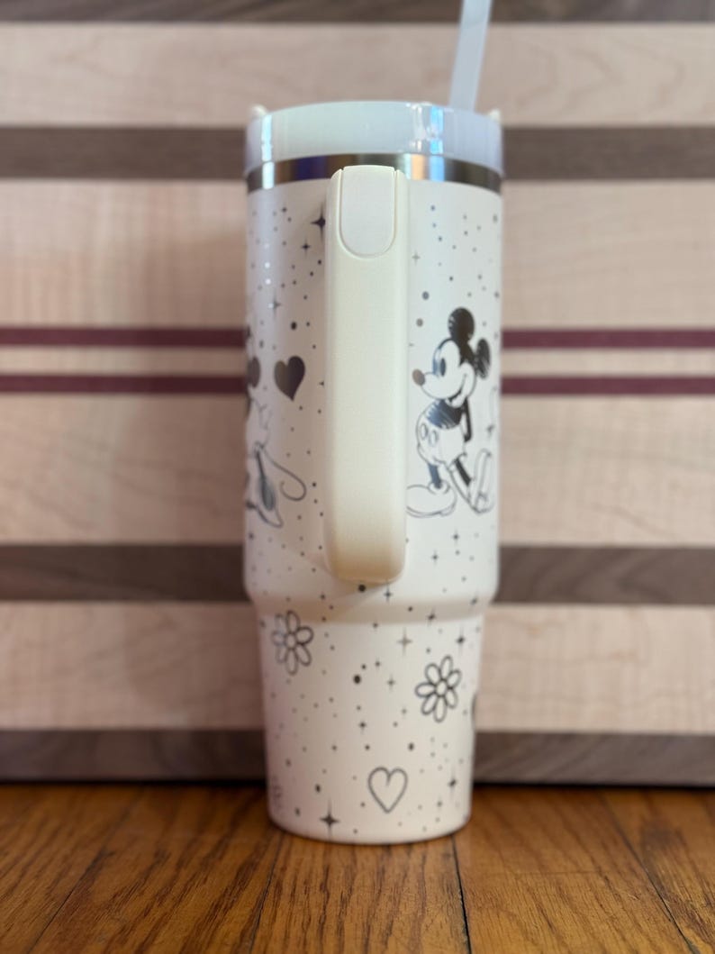 May include: White stainless steel tumbler with a black and white Mickey Mouse and Minnie Mouse design. The tumbler has a black lid and a black handle.
