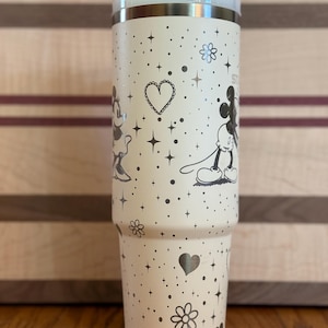 May include: A white stainless steel tumbler with a black and white Mickey Mouse and Minnie Mouse design. The tumbler has a straw and is decorated with hearts, stars, and flowers.