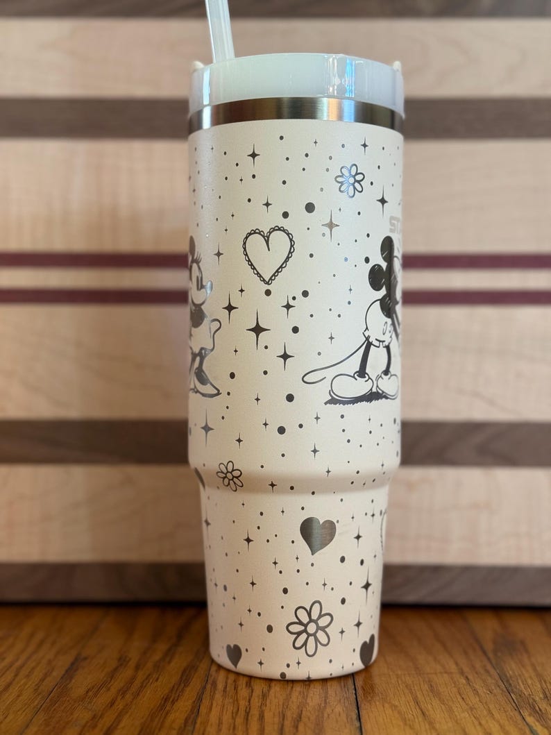 May include: A white stainless steel tumbler with a black and white Mickey Mouse and Minnie Mouse design. The tumbler has a straw and is decorated with hearts, stars, and flowers.