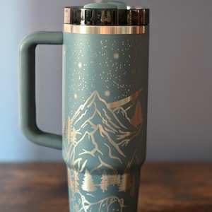 May include: A blue stainless steel travel mug with a black lid and handle. The mug features a laser-etched design of mountains, stars, and bears.