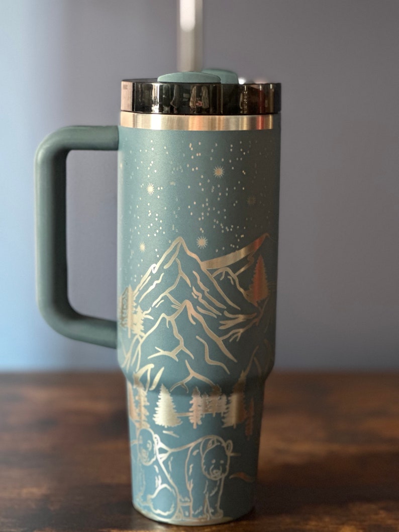 May include: A blue stainless steel travel mug with a black lid and handle. The mug features a laser-etched design of mountains, stars, and bears.
