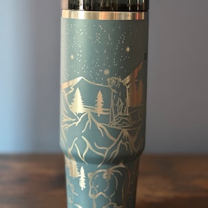 May include: A blue stainless steel tumbler with a gold-colored design of mountains, trees, and bears. The tumbler has a lid with a straw hole.