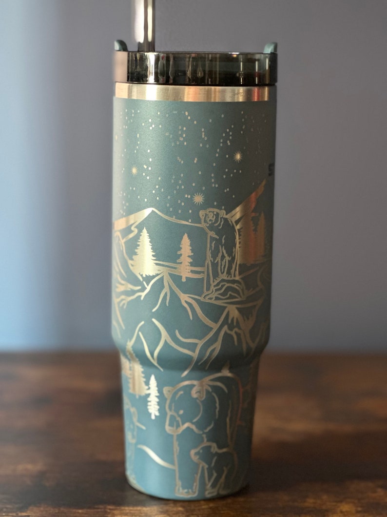 May include: A blue stainless steel tumbler with a gold-colored design of mountains, trees, and bears. The tumbler has a lid with a straw hole.