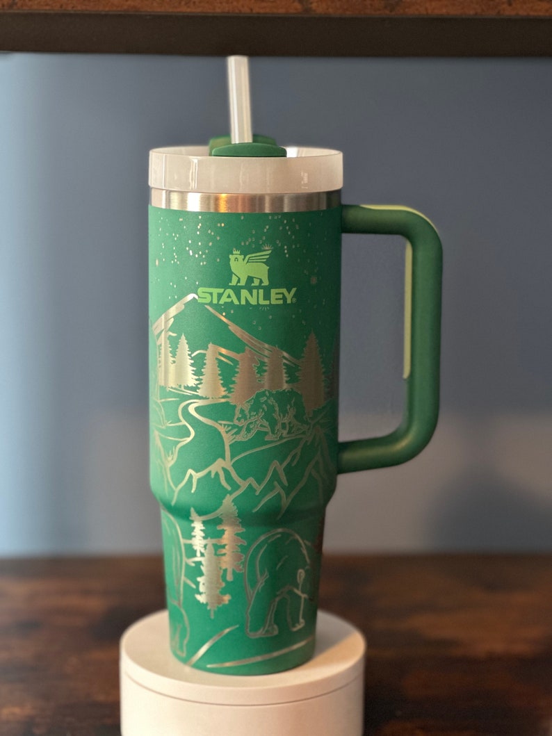 May include: Green stainless steel travel mug with a handle and a straw. The mug has a mountain scene with a bear and trees engraved on it. The mug has the word "STANLEY" on it.