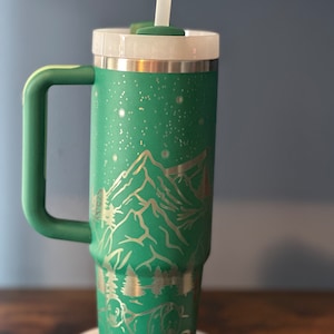 May include: A green stainless steel tumbler with a mountain and bear design. The tumbler has a lid and a straw.