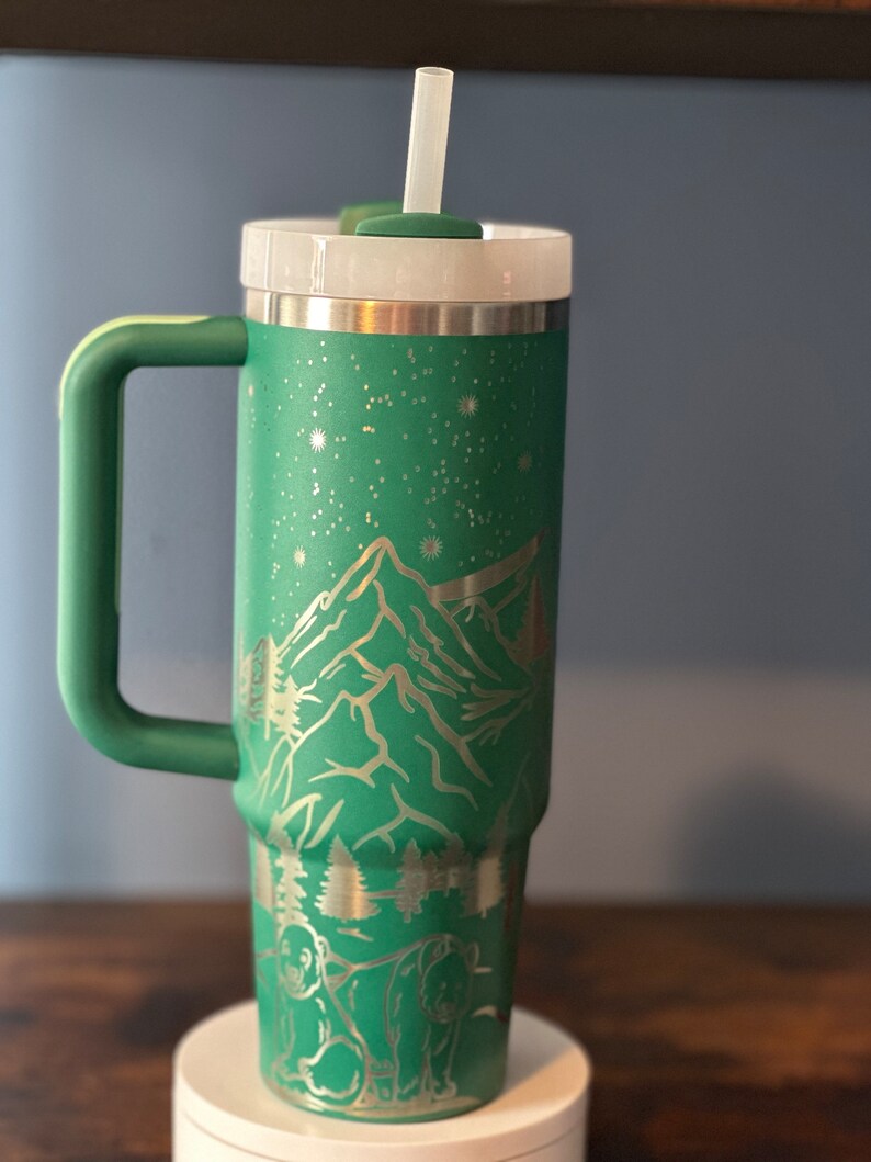 May include: A green stainless steel tumbler with a mountain and bear design. The tumbler has a lid and a straw.