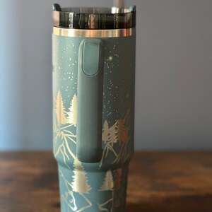 May include: A blue stainless steel travel mug with a black lid and a handle. The mug has a gold design of trees and mountains on it.