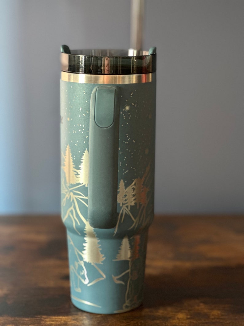 May include: A blue stainless steel travel mug with a black lid and a handle. The mug has a gold design of trees and mountains on it.