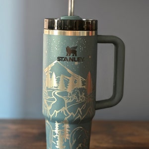 May include: A green stainless steel travel mug with a black lid and a handle. The mug has a gold mountain scene with trees and bears etched into the side. The mug has the word "STANLEY" printed on the side.