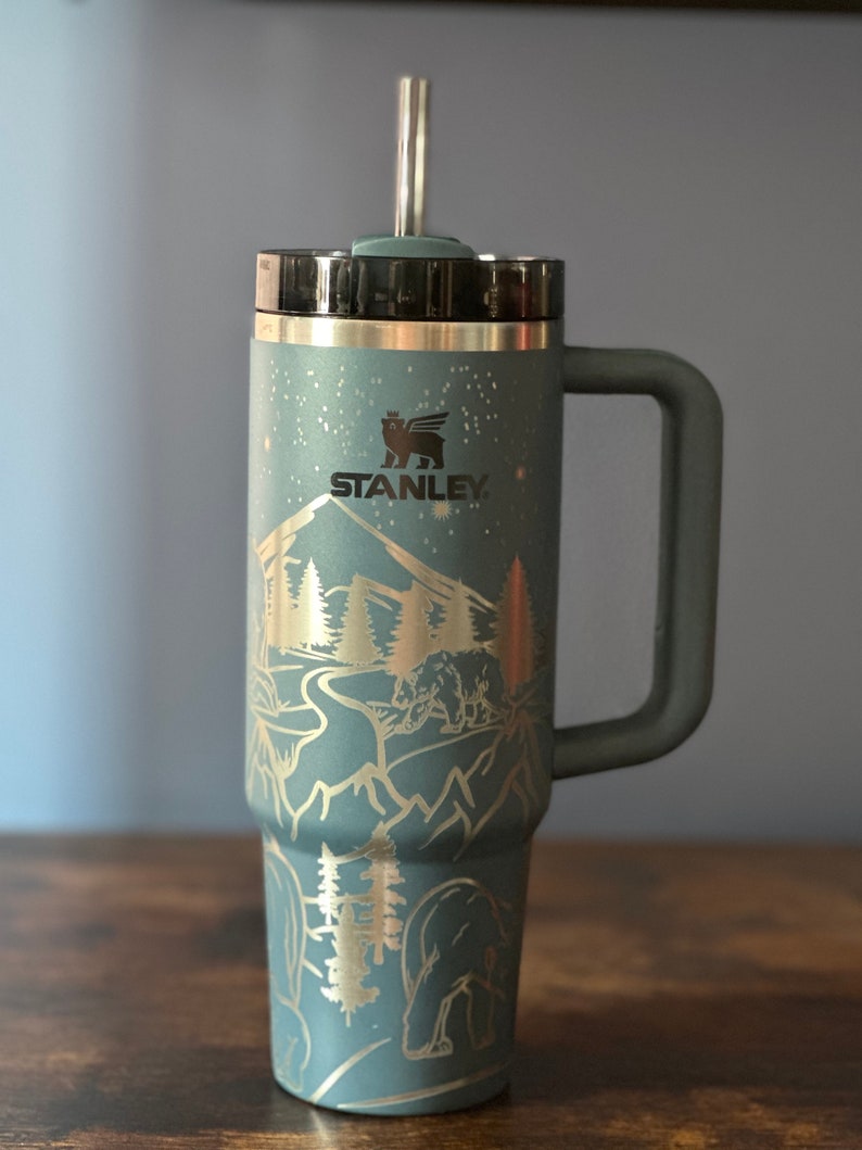 May include: A green stainless steel travel mug with a black lid and a handle. The mug has a gold mountain scene with trees and bears etched into the side. The mug has the word "STANLEY" printed on the side.