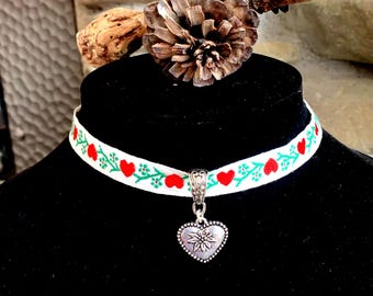 Könnte beinhalten: Weißes Band-Choker mit roten Herzen und grünen Blättern, mit einem silbernen Herz-Anhänger mit Edelweiß-Design.