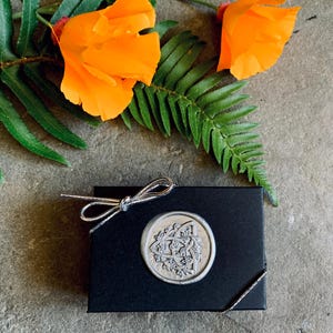 Pode incluir: Uma caixa de presente preta com um la&ccedil;o prateado e um selo de cera decorativo. O selo apresenta um design detalhado em forma circular. Flores laranja brilhantes e folhagem verde s&atilde;o dispostas ao redor da caixa sobre uma superf&iacute;cie de pedra.