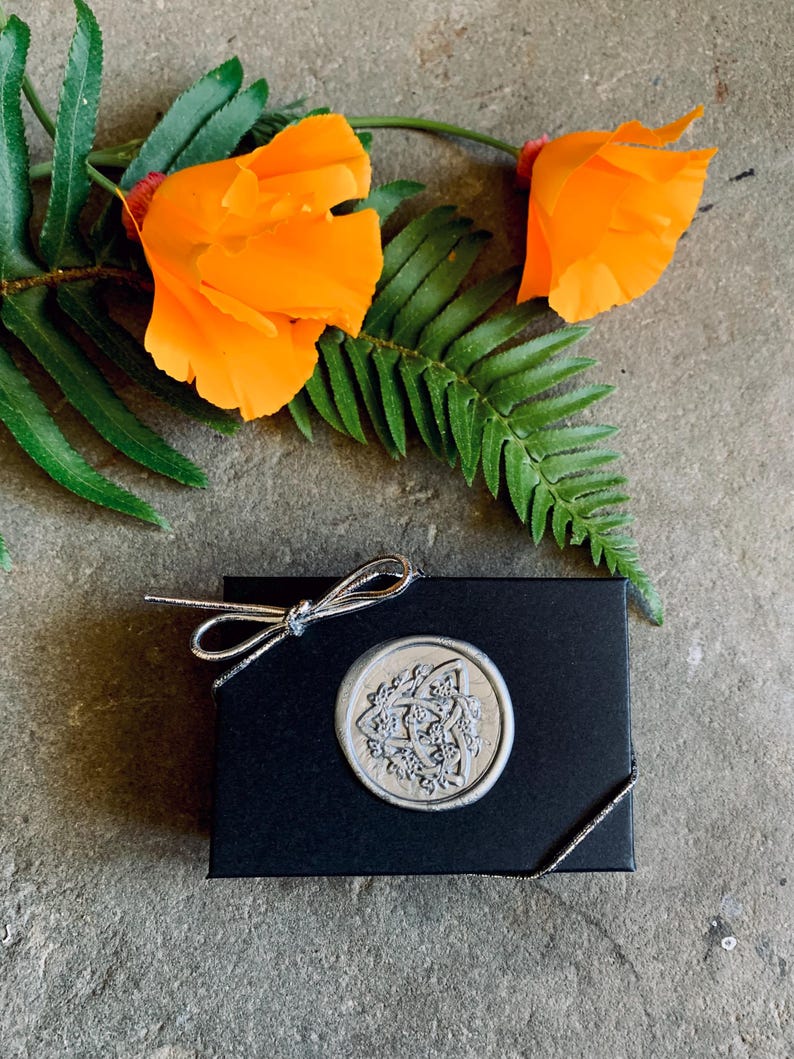 Pode incluir: Uma caixa de presente preta com um la&ccedil;o prateado e um selo de cera decorativo. O selo apresenta um design detalhado em forma circular. Flores laranja brilhantes e folhagem verde s&atilde;o dispostas ao redor da caixa sobre uma superf&iacute;cie de pedra.