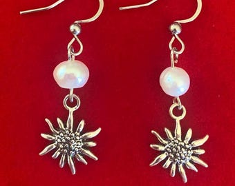 May include: A pair of silver-toned earrings with a sunflower design. Each earring features a white pearl bead and a detailed sunflower charm. The earrings hang from a simple hook, set against a red background.