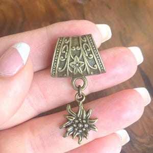 Puede incluir: Un colgante de color bronce con una parte superior rectangular adornada y un dije de flor estilizada colgante. La parte superior presenta diseños florales y de volutas. El colgante se sostiene en una mano, mostrando sus intrincados detalles y estética vintage.