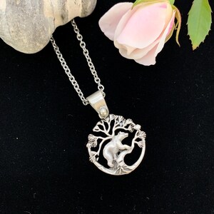 Pode incluir: Colar com pingente prateado com um urso escalando uma &aacute;rvore dentro de uma moldura circular. O pingente pende de uma corrente delicada. Uma rosa rosa e uma pedra cinza est&atilde;o no fundo, em um fundo preto.