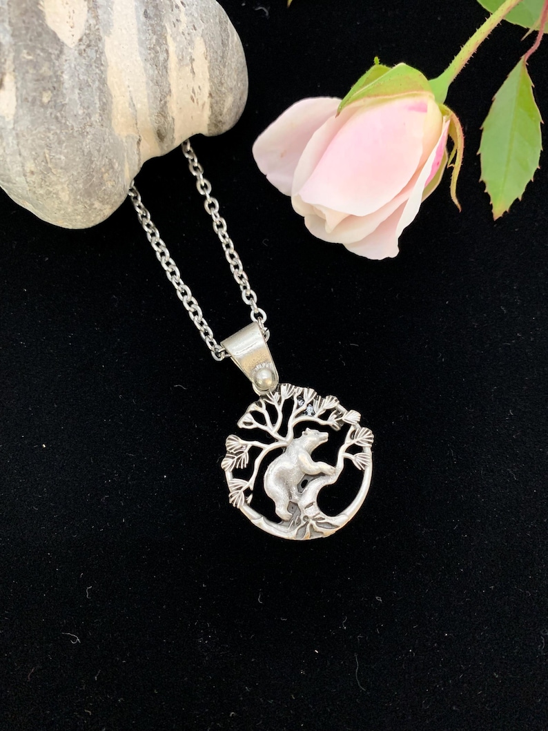 Pode incluir: Colar com pingente prateado com um urso escalando uma &aacute;rvore dentro de uma moldura circular. O pingente pende de uma corrente delicada. Uma rosa rosa e uma pedra cinza est&atilde;o no fundo, em um fundo preto.