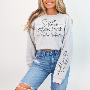 May include: A gray cropped sweatshirt with the text "Surround yourself with Positive Vibes" and a small floral design. The sweatshirt has a long sleeve with the text "and watch your life transform" on the cuff.