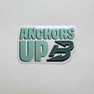 May include: A rectangular sticker with the words "ANCHORS UP" in a teal and black color scheme. The "B" in "UP" is stylized with a wave pattern. The sticker is on a white background.