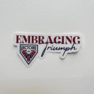 May include: A white sticker with the words "EMBRACING triumph" in a mix of burgundy and navy blue script. The word "VICTOIRE" is in a diamond-shaped emblem with burgundy and navy blue accents.
