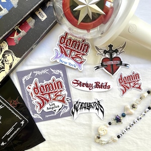May include: An assortment of stickers and accessories featuring the band Stray Kids. Stickers include the band's logo and city names like New York and Los Angeles. A light stick and beaded necklace are also visible.